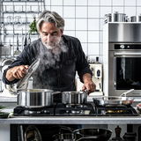 Mann in moderner Küche hebt den Deckel eines dampfenden Topfes an, umgeben von einem hochwertigen 4-teiligen Topfset aus Edelstahl auf einem Herd.