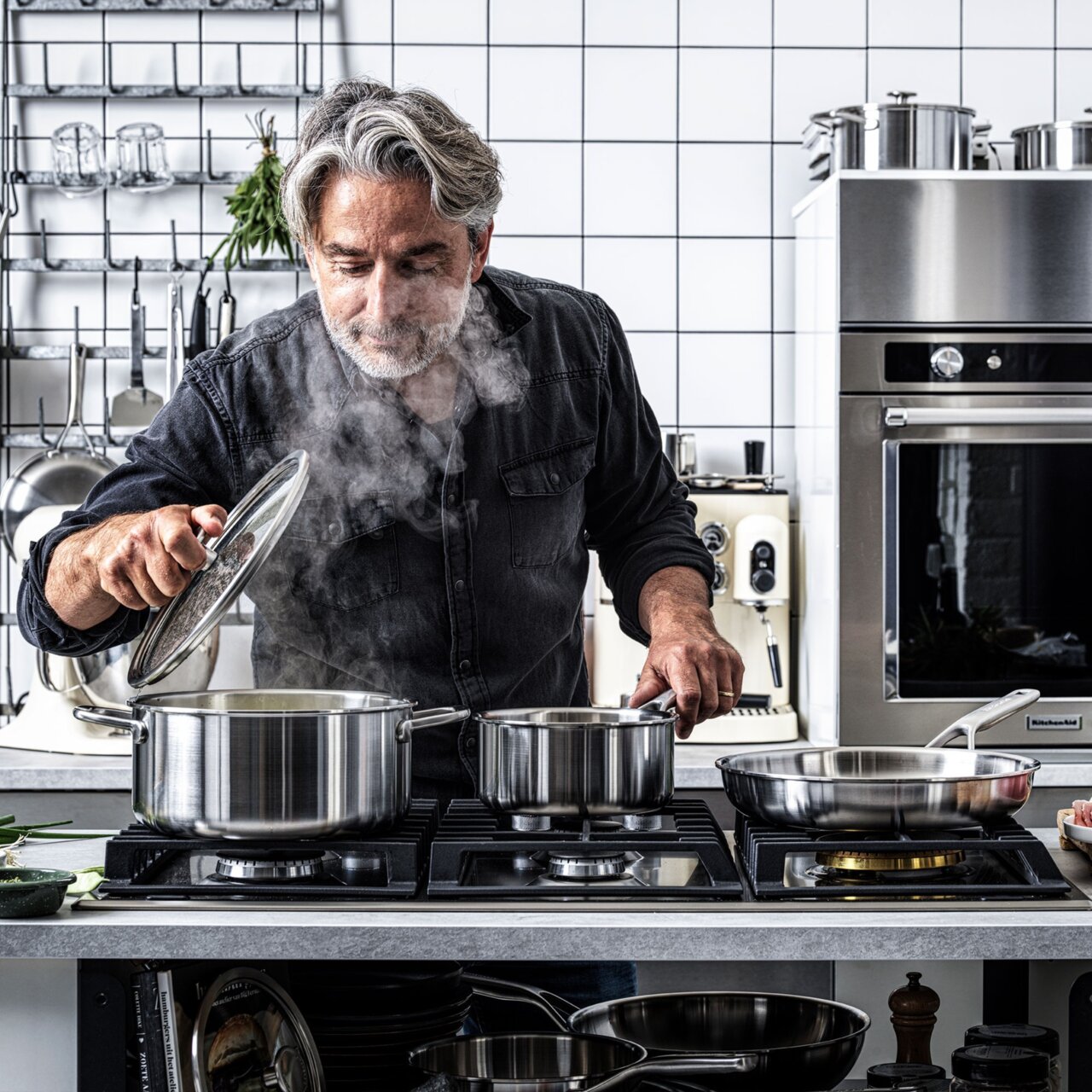 Mann in moderner Küche hebt den Deckel eines dampfenden Topfes an, umgeben von einem hochwertigen 4-teiligen Topfset aus Edelstahl auf einem Herd.