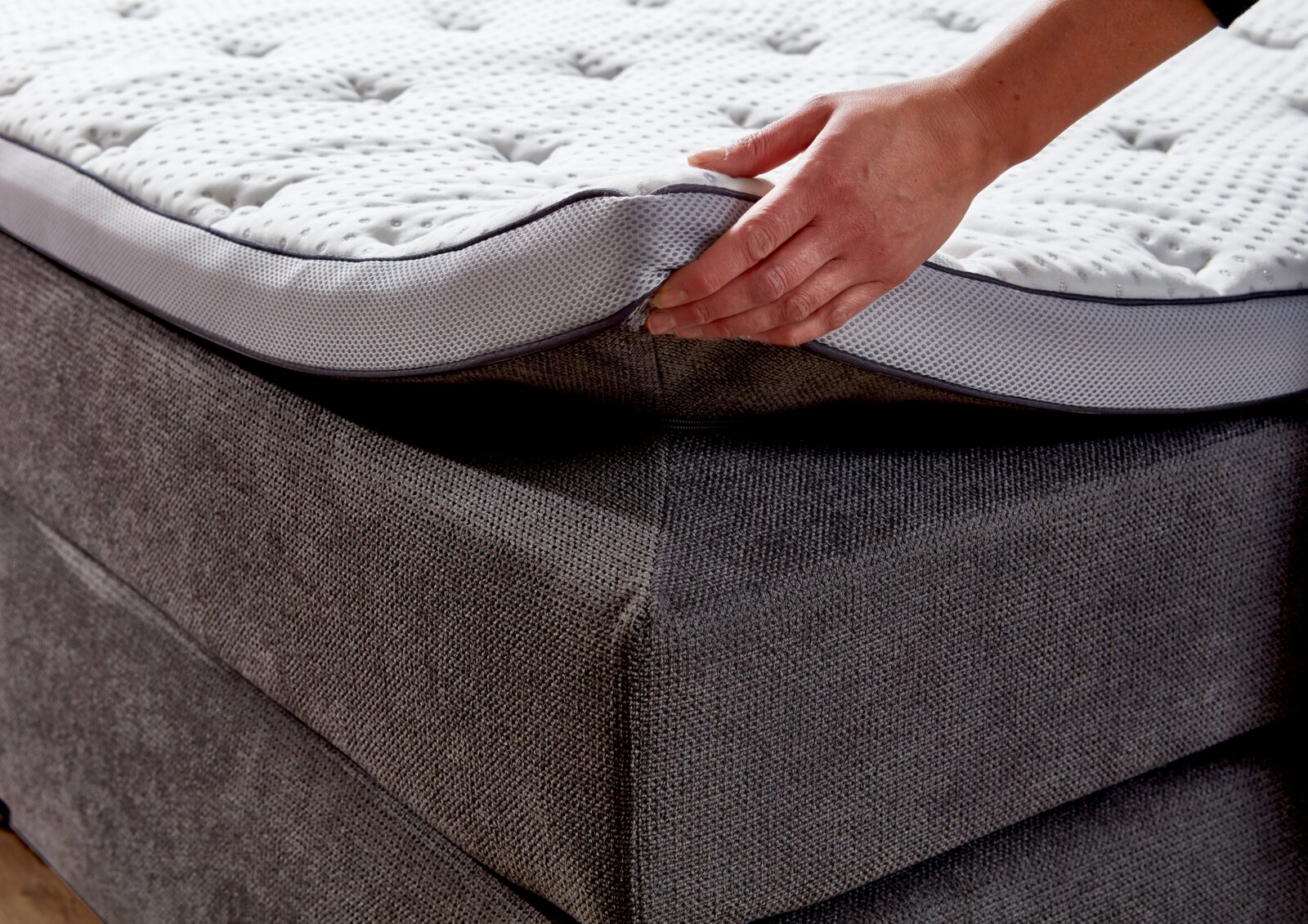 Close-up van een boxspringbed met een hand die het matras optilt om de constructie en de bekleding te laten zien.