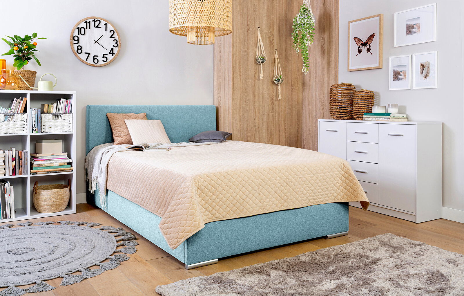 Gemütliches Schlafzimmer mit einem Boxspringbett in hellblauem Stoffbezug, umgeben von moderner Dekoration und Möbeln, aus seitlicher Perspektive.