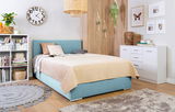Gemütliches Schlafzimmer mit einem Boxspringbett in hellblauem Stoffbezug, umgeben von moderner Dekoration und Möbeln, aus seitlicher Perspektive.