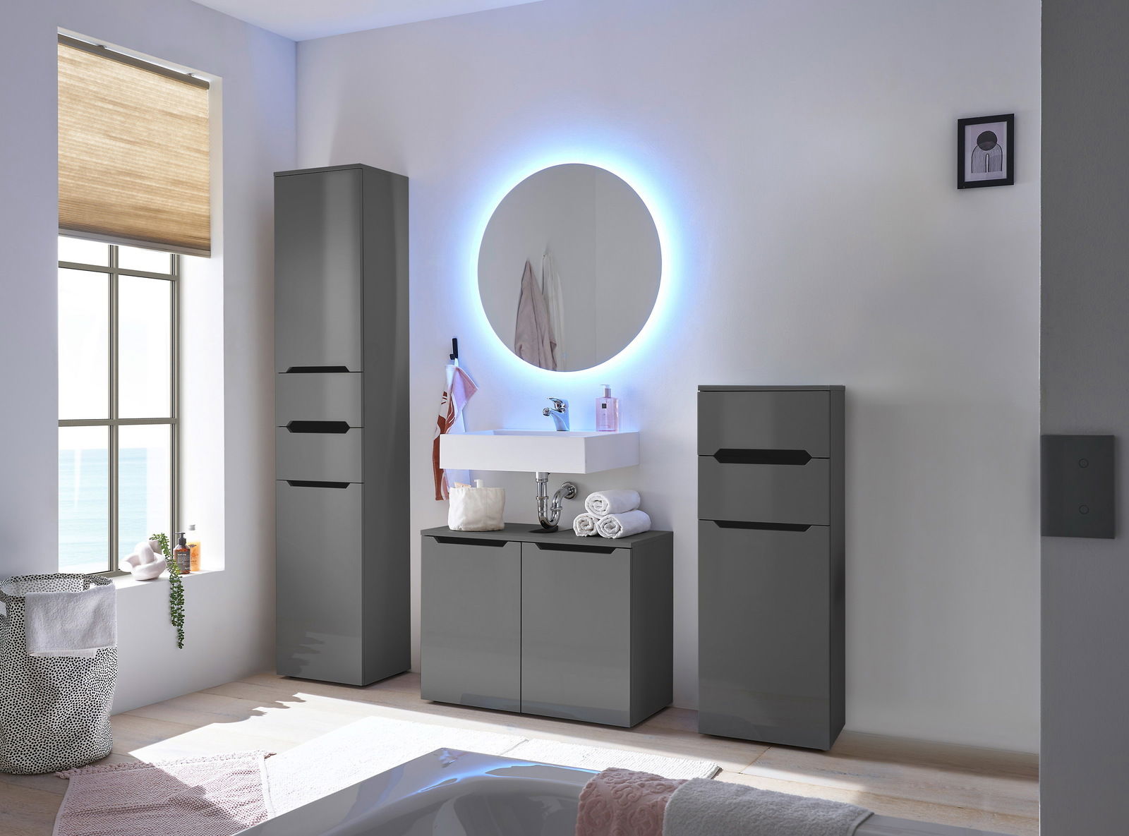 Moderne Hochkommode in einem stilvollen Badezimmer, frontal betrachtet. Die Kommode ist in einem eleganten, dunklen Farbton gehalten und steht neben einem beleuchteten Spiegel und einem Waschbecken.