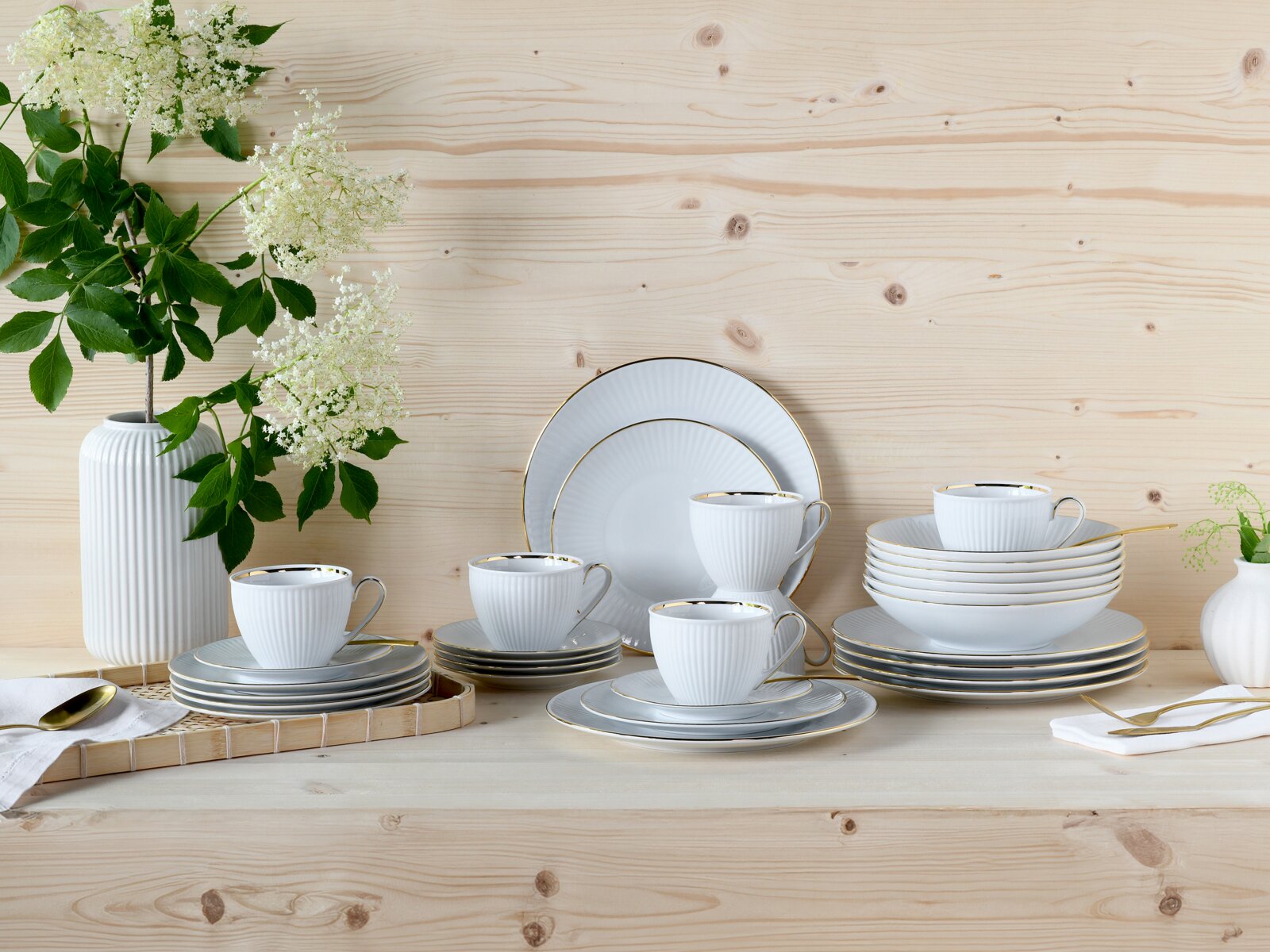 Creatable Kombiservice 30-tlg VALENCIA Elegantes weißes Porzellan-Geschirrset mit feiner Rillenstruktur und goldfarbenem Rand, mit Tassen, Tellern und Schalen auf hellem Holztisch arrangiert, dekoriert mit Vase und weißen Blüten, Perspektive: frontale leicht erhöhte Ansicht.