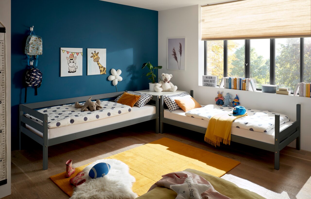 Kinderzimmer mit zwei grauen Etagenbetten in L-Form, dekoriert mit bunten Kissen und Spielzeug, Blick aus dem Fenster auf einen sonnigen Garten.