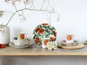 Blumig gemustertes Keramik-Geschirrset in Weiß mit orangefarbenen Blüten und grünen Blättern, bestehend aus Tellern, Schalen und Tassen, auf einem Holztisch arrangiert; Perspektive: frontale Ansicht auf Augenhöhe.