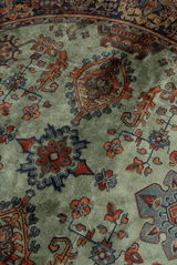 Nahaufnahme eines Teppichs mit traditionellem Muster in Rot- und Blautönen auf grünem Hintergrund, aus schräger Perspektive fotografiert.