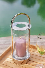 Elegantes Windlicht mit Glas und brennender Kerze, auf einem Holztisch neben einem Glas Weißwein, mit Blick auf einen See im Hintergrund.