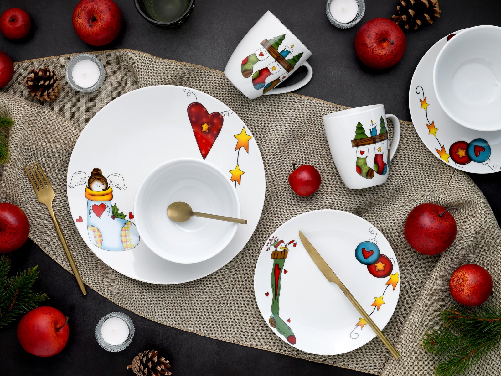 Draufsicht auf ein festlich dekoriertes Porzellan-Geschirrset in Weiß mit weihnachtlichen Motiven (Herzen, Sterne, Engel und Strümpfe), bestehend aus Teller, Schalen und Tassen, arrangiert auf Jute mit goldfarbenem Besteck, roten Äpfeln, Tannenzapfen und