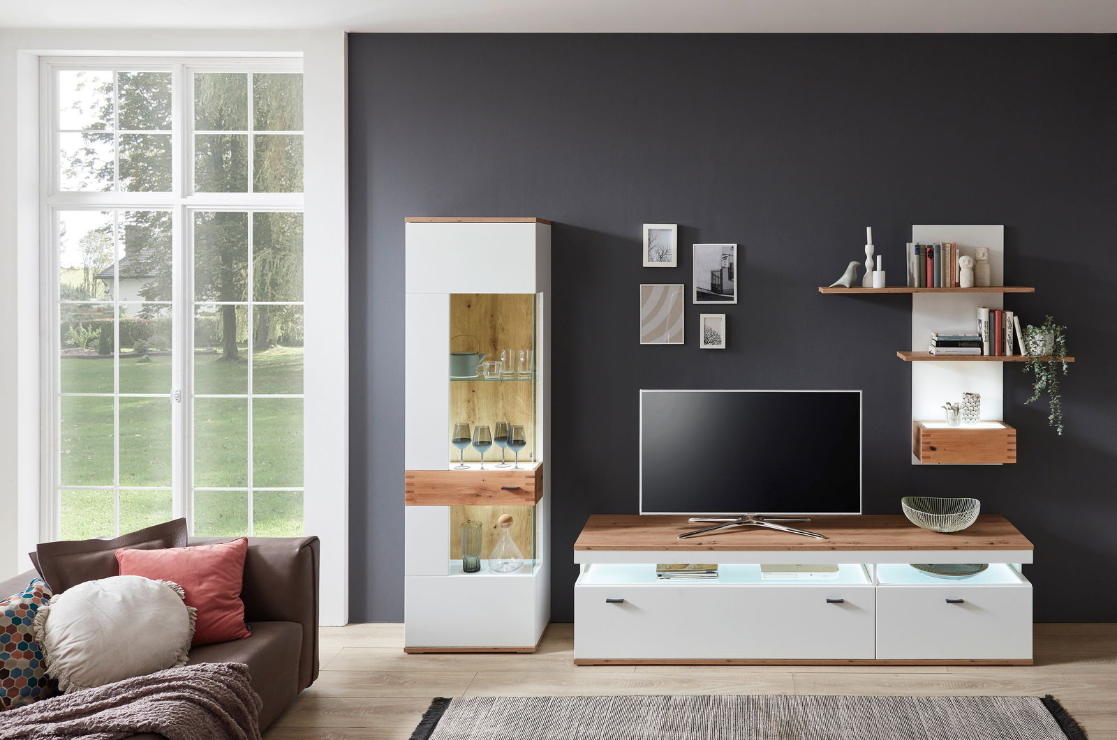 Moderne Wandkombination in einem Wohnzimmer mit weißem und holzfarbenem Design, bestehend aus einem TV-Schrank, einem hohen Vitrinenschrank und Wandregalen, aus der Perspektive von vorne betrachtet.