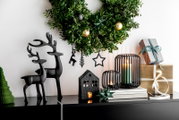 Frontalansicht einer stilvollen Weihnachtsdekoration mit grünen Kränzen, schwarzen Rentierfiguren, leuchtenden Windlichtern und festlich verpackten Geschenken auf einem Sideboard.