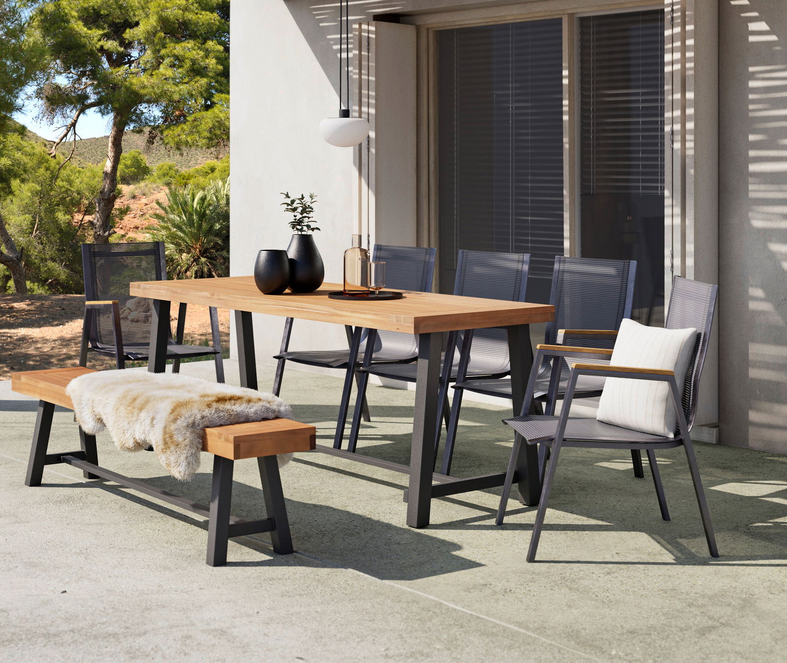 Moderner Gartentisch mit Holzplatte und Metallgestell, umgeben von stilvollen Stühlen und einer Bank mit Fellauflage, in einer sonnigen Terrassenumgebung.