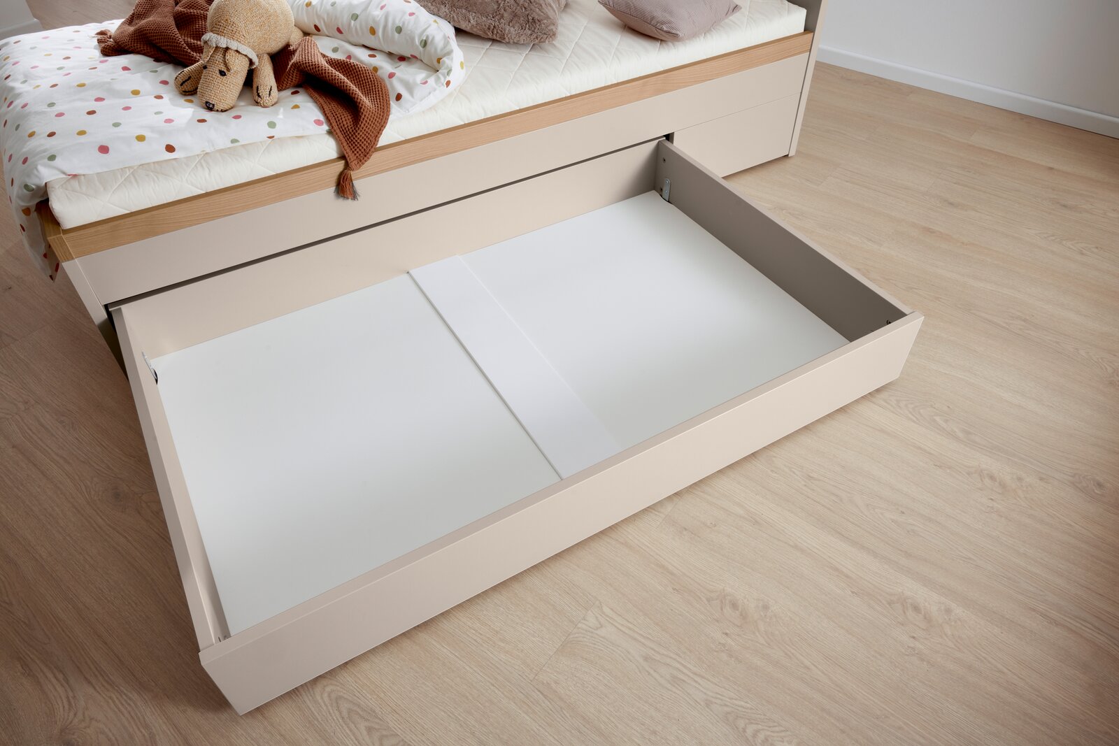 Bettkasten in hellem Holzton, von oben fotografiert, teilweise unter einem Bett hervorgezogen, mit Bettzeug und Kuscheltier auf dem Bett.