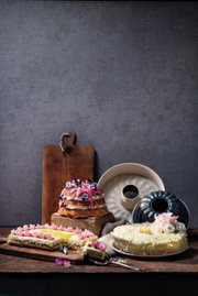 Verschiedene Kuchen auf einem rustikalen Holztisch, darunter ein rechteckiger Kuchen mit rosa Dekoration und ein runder Kuchen mit weißen Blumen. Im Hintergrund sind zwei Gugelhupfformen zu sehen. Perspektive: seitlich.