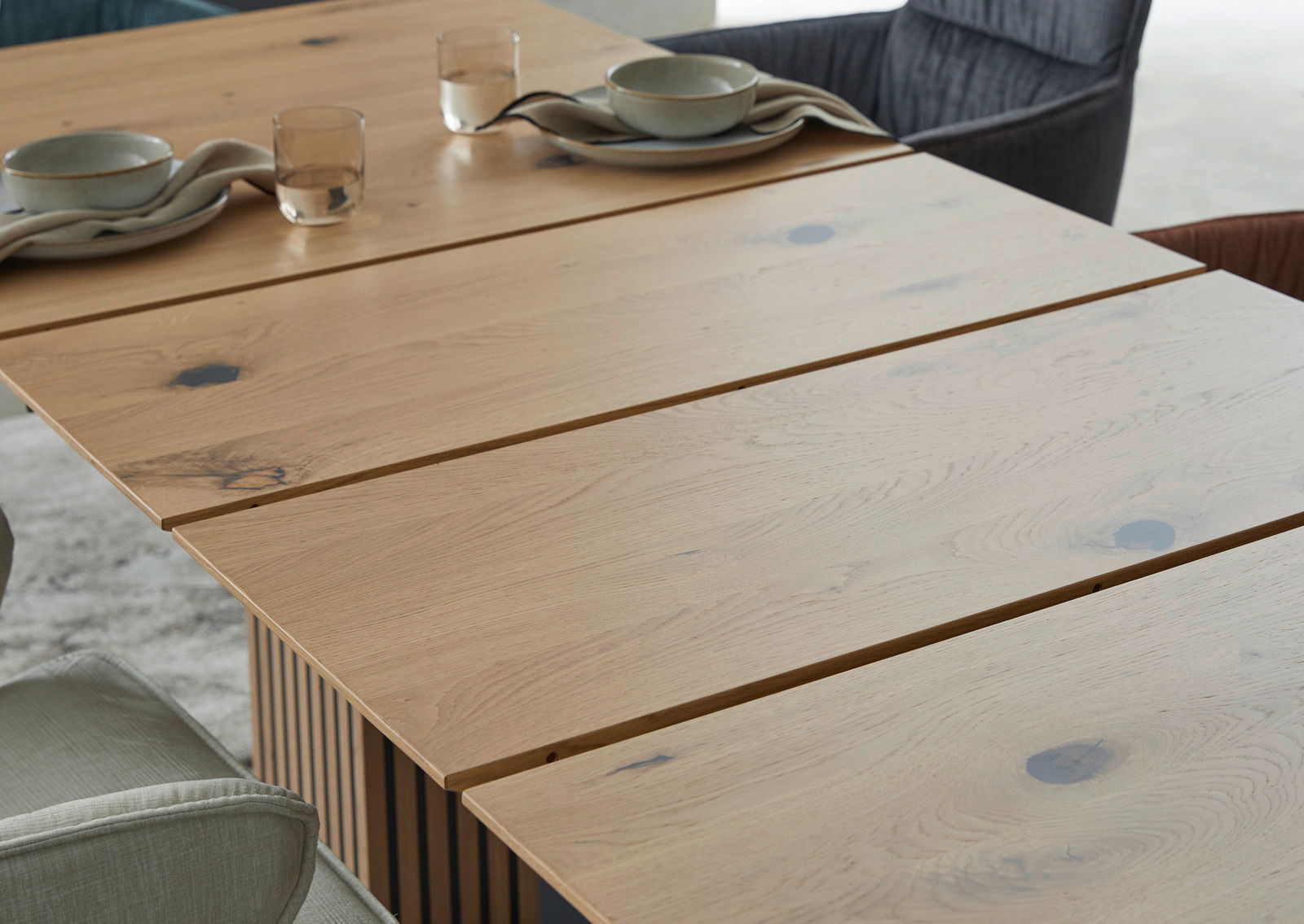 Nahaufnahme eines Esstisches aus hellem Holz mit natürlicher Maserung, gedeckt mit Geschirr und Gläsern, aus seitlicher Perspektive.