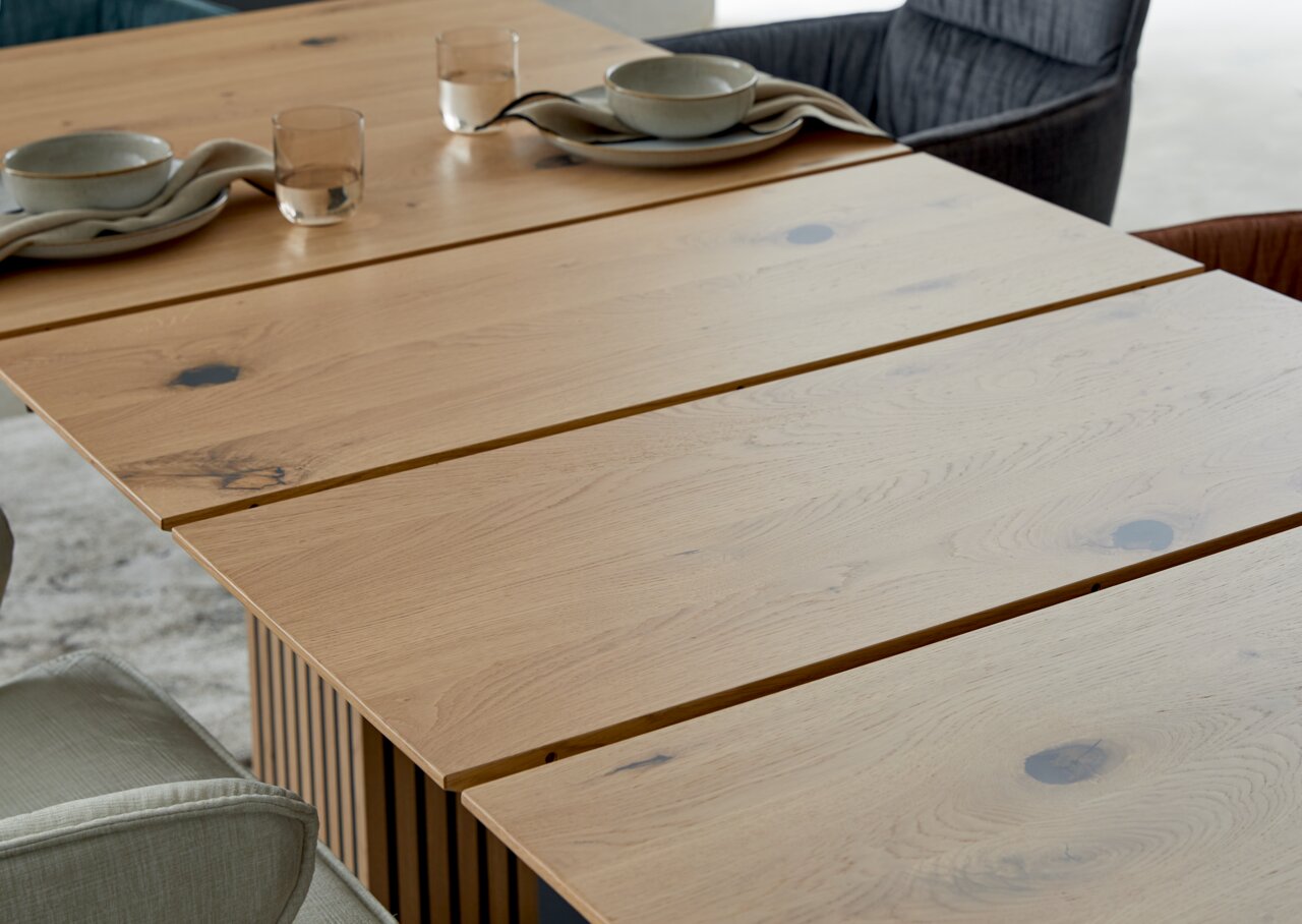 Nahaufnahme eines Esstisches aus hellem Holz mit natürlicher Maserung, gedeckt mit Geschirr und Gläsern, aus seitlicher Perspektive.