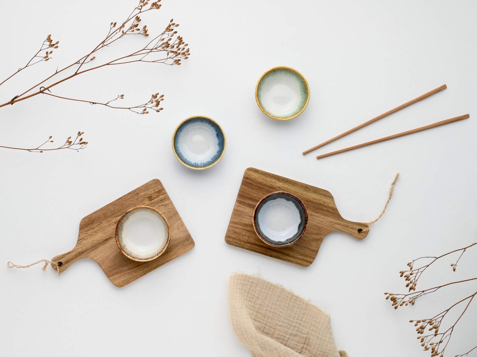 Flatlay in Draufsicht: vier kleine glasierte Keramikschälchen in Blau-, Grün- und Cremetönen auf zwei Holz-Servierbrettern, daneben Holzstäbchen und dekorative Trockenblumen auf weißem Hintergrund