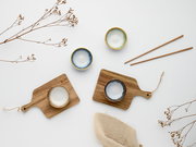 Flatlay in Draufsicht: vier kleine glasierte Keramikschälchen in Blau-, Grün- und Cremetönen auf zwei Holz-Servierbrettern, daneben Holzstäbchen und dekorative Trockenblumen auf weißem Hintergrund