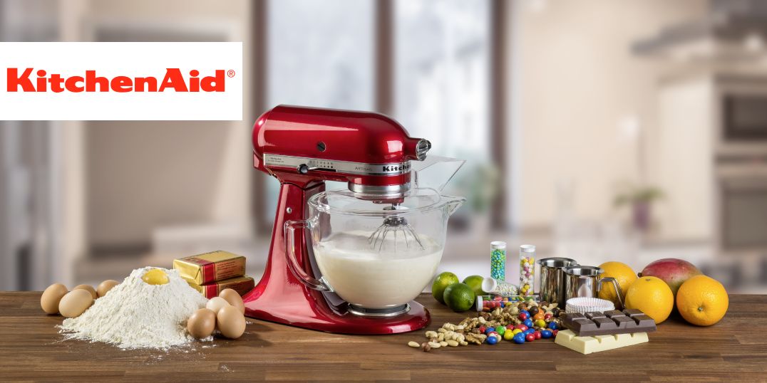 Ein roter KitchenAid-Mixer mit einer Glasschüssel, umgeben Backzubehör auf einer Holzarbeitsplatte.