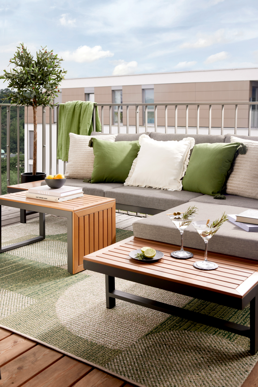 Moderne Gartenlounge auf einem Balkon mit grauem Sofa, grünen und weißen Kissen, Holztischen und dekorativen Elementen, seitliche Perspektive