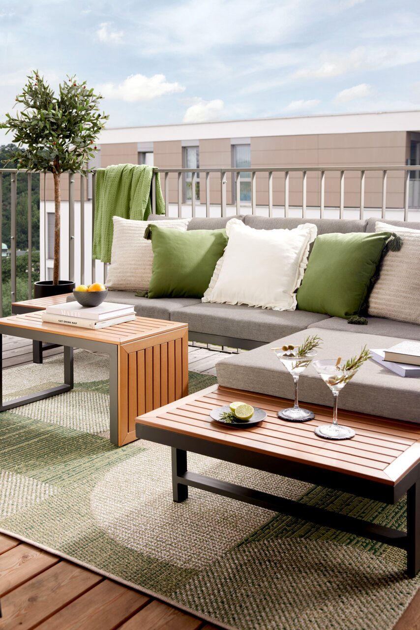 Moderne tuinlounge op een balkon met een grijze bank, groene en witte kussens, houten tafels en decoratieve elementen, zijaanzicht