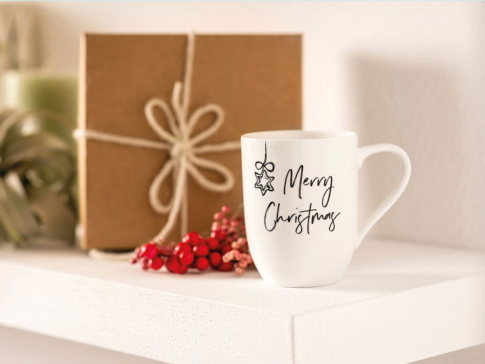 Stimmungsvolle Weihnachtsdekoration mit weißer „Merry Christmas“-Tasse, Geschenk in Naturpapier und roten Beeren auf einer hellen Ablage