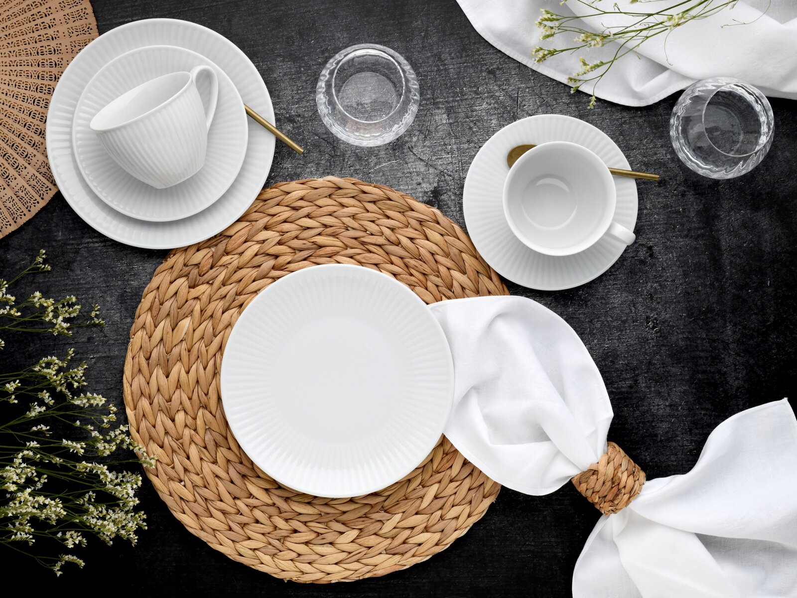 Draufsicht auf ein stilvoll gedecktes Tischarrangement mit weißem geriffeltem Porzellan-Geschirr (Teller, Schalen, Tassen), goldfarbenen Löffeln, geflochtenen Natur-Platzsets und weißen Servietten auf dunkler Tischplatte, ergänzt durch Glas-Teelichthalter