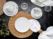 Draufsicht auf ein stilvoll gedecktes Tischarrangement mit weißem geriffeltem Porzellan-Geschirr (Teller, Schalen, Tassen), goldfarbenen Löffeln, geflochtenen Natur-Platzsets und weißen Servietten auf dunkler Tischplatte, ergänzt durch Glas-Teelichthalter