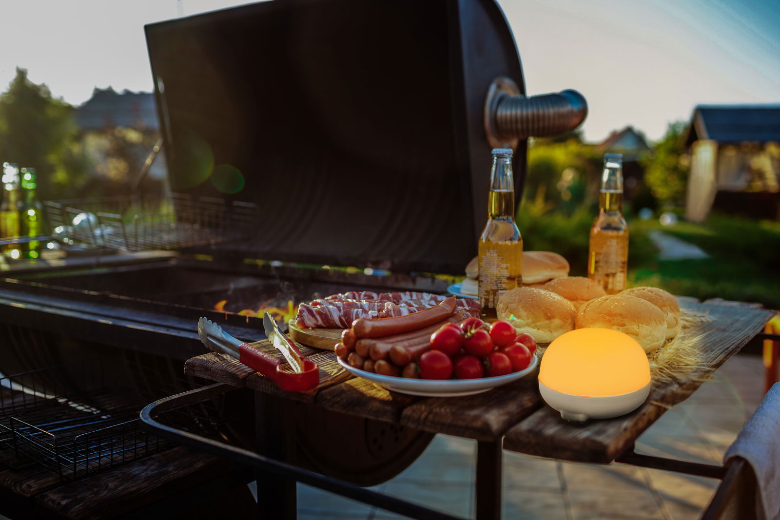 Akku-Leuchtmittel auf einem Tisch neben einem Grill, umgeben von Grillgut und Getränken, bei Sonnenuntergang im Freien.