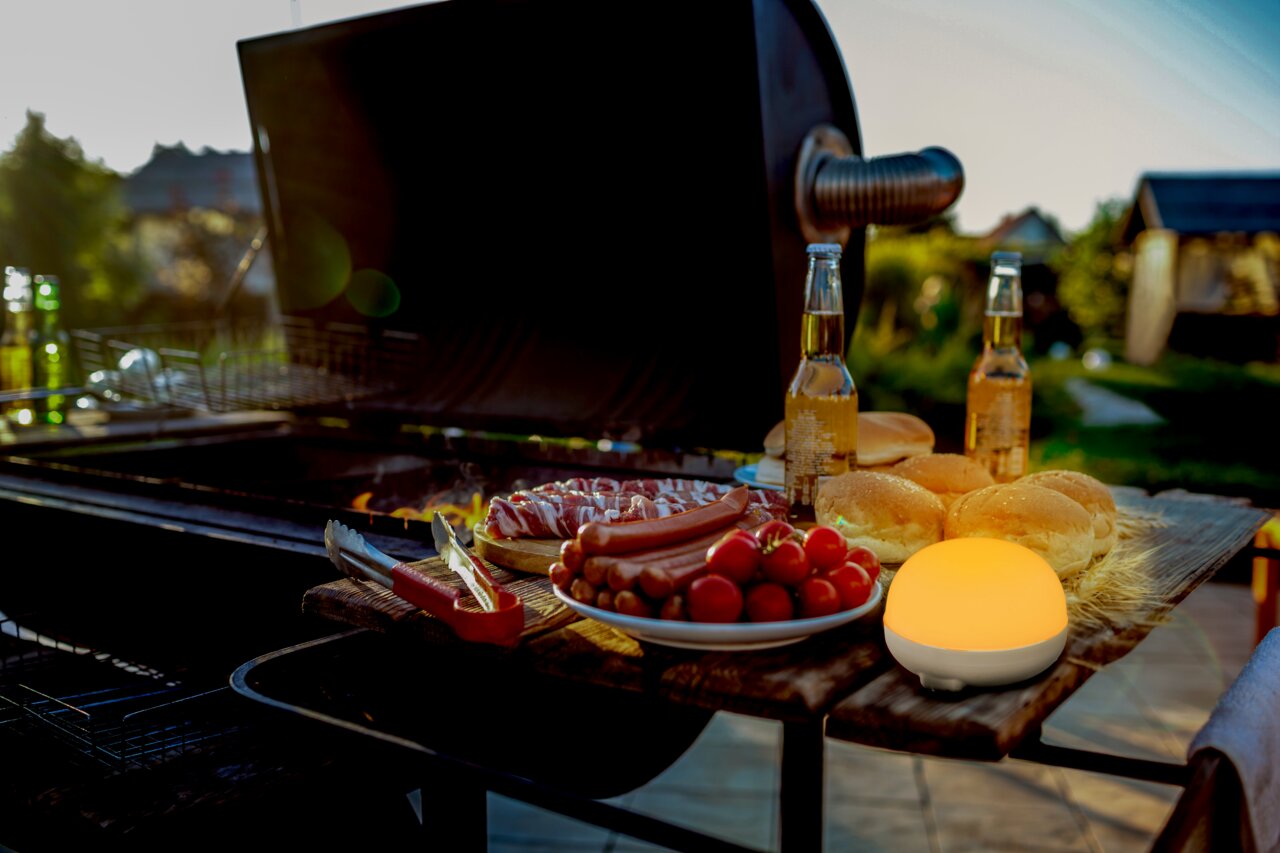 Akku-Leuchtmittel auf einem Tisch neben einem Grill, umgeben von Grillgut und Getränken, bei Sonnenuntergang im Freien.