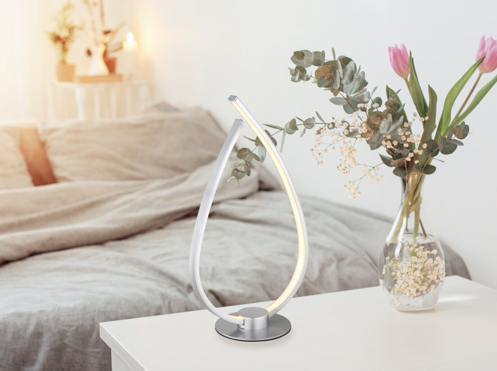 Moderne silberne LED-Tischlampe in geschwungener Tropfenform mit warmweißem Licht auf weißem Nachttisch im Schlafzimmer, Perspektive: leicht schräg von vorn