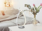 Moderne silberne LED-Tischlampe in geschwungener Tropfenform mit warmweißem Licht auf weißem Nachttisch im Schlafzimmer, Perspektive: leicht schräg von vorn