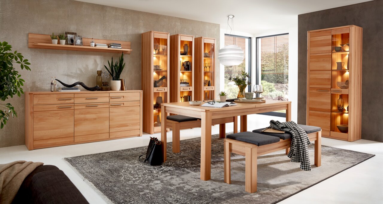 Wohnzimmer mit modernen Holzmöbeln, darunter ein Sideboard, Vitrinen und ein Esstisch mit Bänken, aus einer frontalen Perspektive fotografiert.