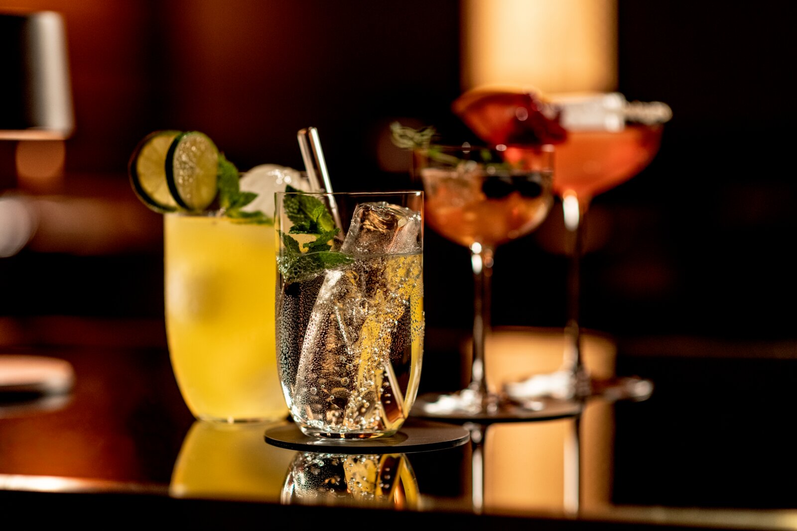 Nahaufnahme auf Augenhöhe von drei Cocktails auf einem glänzenden Bartresen: vorn ein sprudelnder Drink im Tumbler mit Eis, Minze und Metallstrohhalm, daneben ein gelber Cocktail mit Limettenscheiben, im Hintergrund rosafarbene Drinks in Stielgläsern.