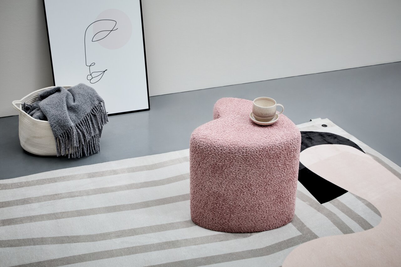 Rosa Hocker in Herzform, von schräg oben fotografiert, mit einer Tasse darauf, auf einem gemusterten Teppich.