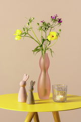 Tischlicht in flieder gesprenkelt auf einem gelben Tisch, neben einer Vase mit Blumen und zwei dekorativen Hasenfiguren. Perspektive: Frontalansicht.