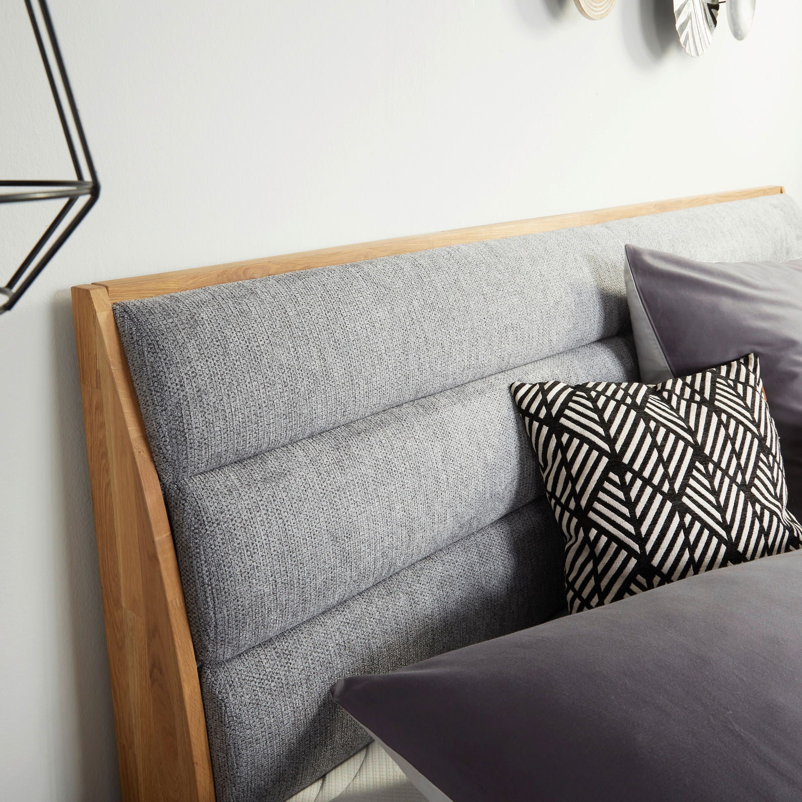 Nahaufnahme des gepolsterten Kopfteils eines Boxspringbetts mit grauem Stoffbezug und Holzelementen, dekoriert mit einem schwarz-weiß gemusterten Kissen, seitliche Perspektive.