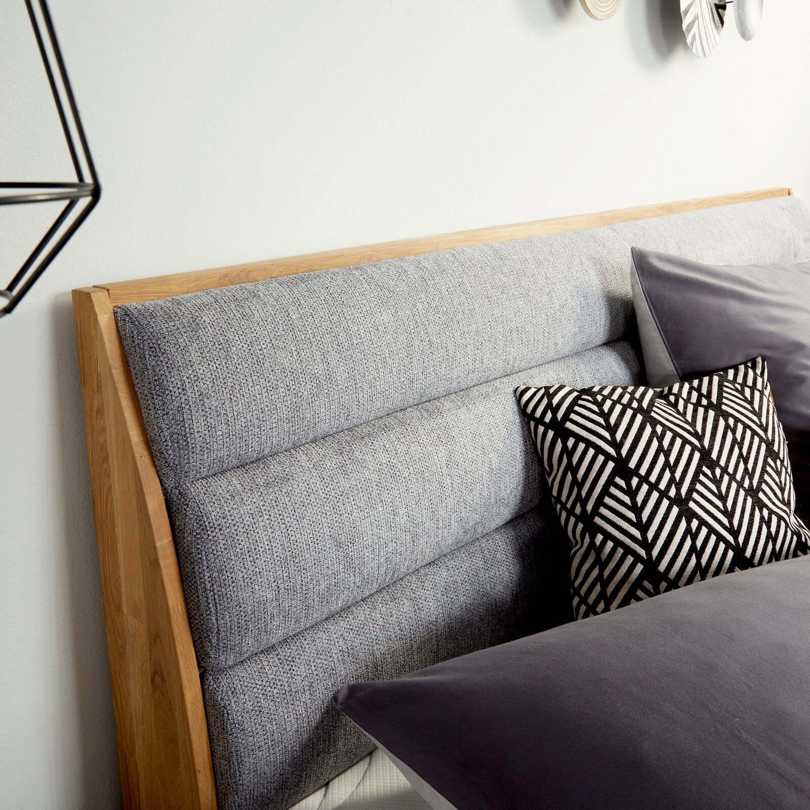 Nahaufnahme des gepolsterten Kopfteils eines Boxspringbetts mit grauem Stoffbezug und Holzelementen, dekoriert mit einem schwarz-weiß gemusterten Kissen, seitliche Perspektive.