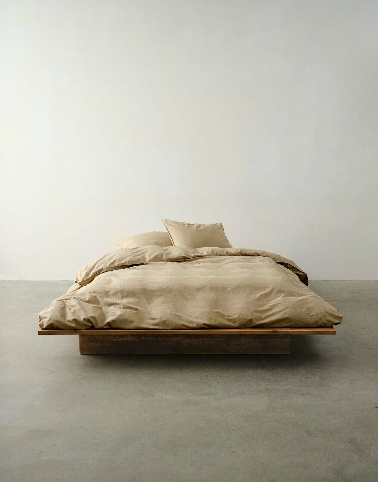 Beige Bettwäsche auf einem niedrigen Holzrahmenbett, fotografiert aus der Vorderansicht in einem minimalistischen Raum.