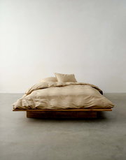 Beige Bettwäsche auf einem niedrigen Holzrahmenbett, fotografiert aus der Vorderansicht in einem minimalistischen Raum.