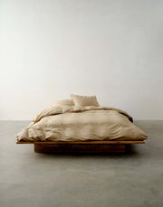 Beige Bettwäsche auf einem niedrigen Holzrahmenbett, fotografiert aus der Vorderansicht in einem minimalistischen Raum.