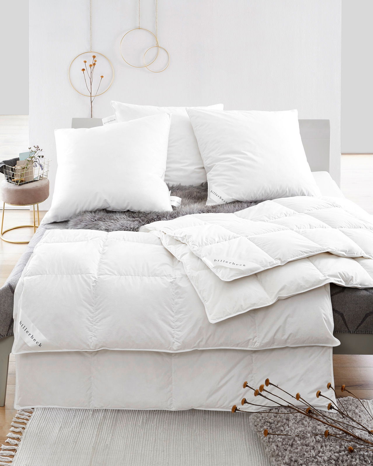 Blick auf ein Bett mit weißen Daunenkissen und Daunendecke, dekoriert mit einem grauen Fellüberwurf.