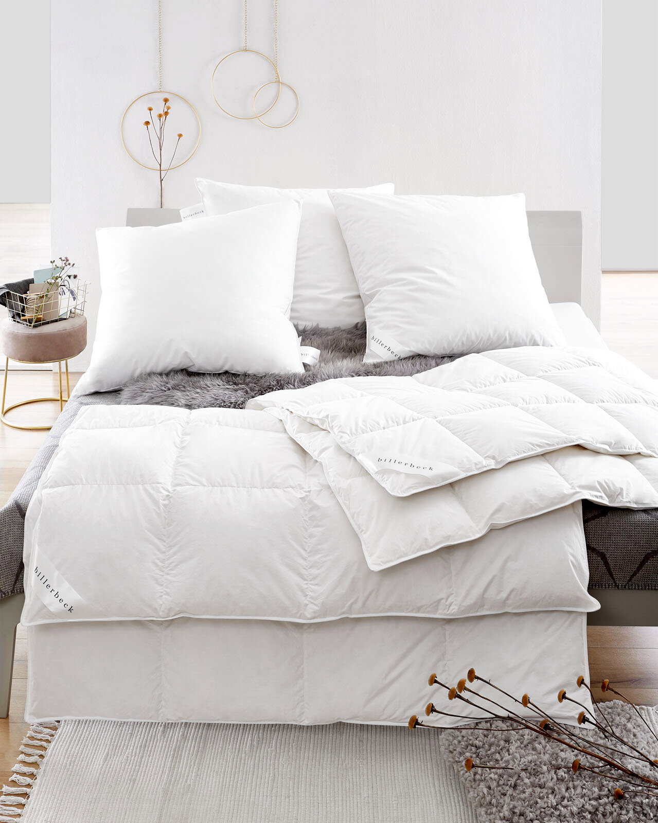 Blick auf ein Bett mit weißen Daunenkissen und Daunendecke, dekoriert mit einem grauen Fellüberwurf.