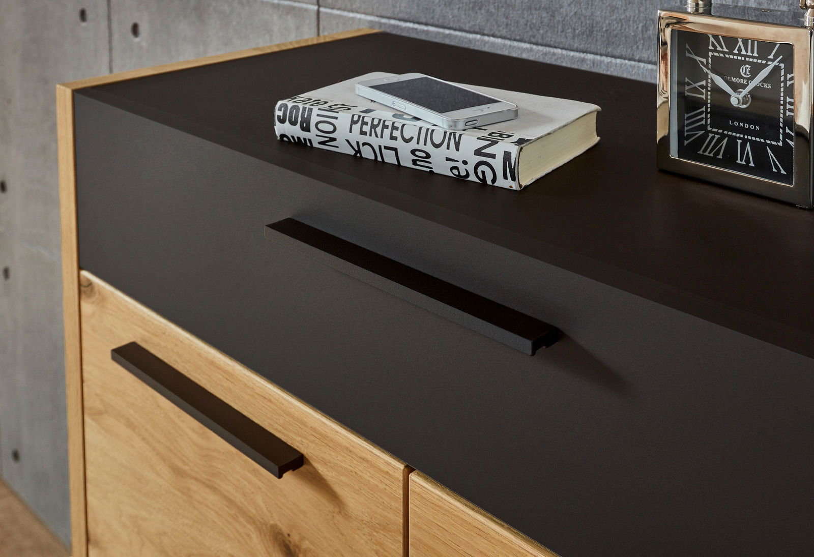 Nahaufnahme einer modernen Kommode mit dunkler Oberfläche und Holzfront, darauf ein Buch, ein Smartphone und eine Uhr. Perspektive von schräg oben.