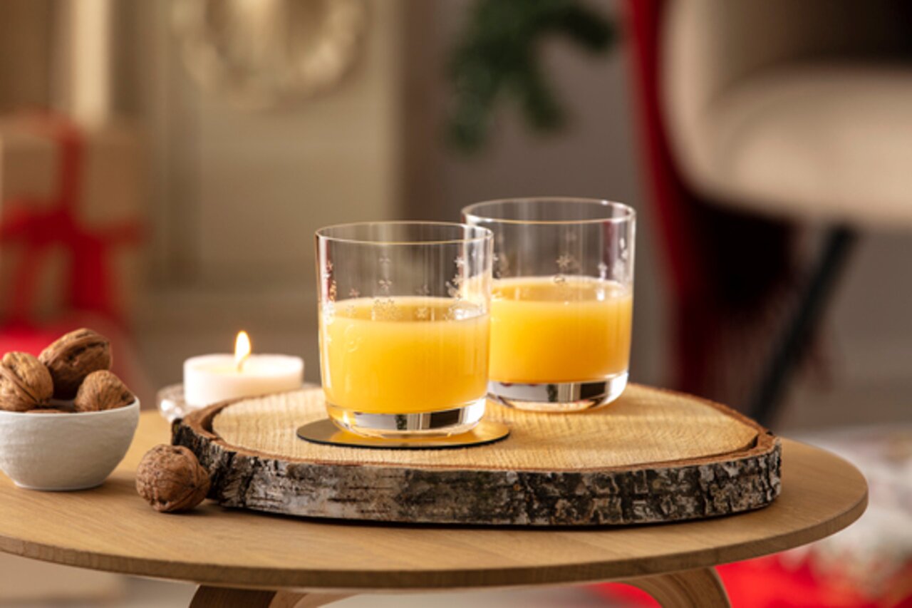 Zwei Wassergläser mit Orangensaft auf einem rustikalen Holztablett, umgeben von Nüssen und einer brennenden Kerze, aus seitlicher Perspektive fotografiert.