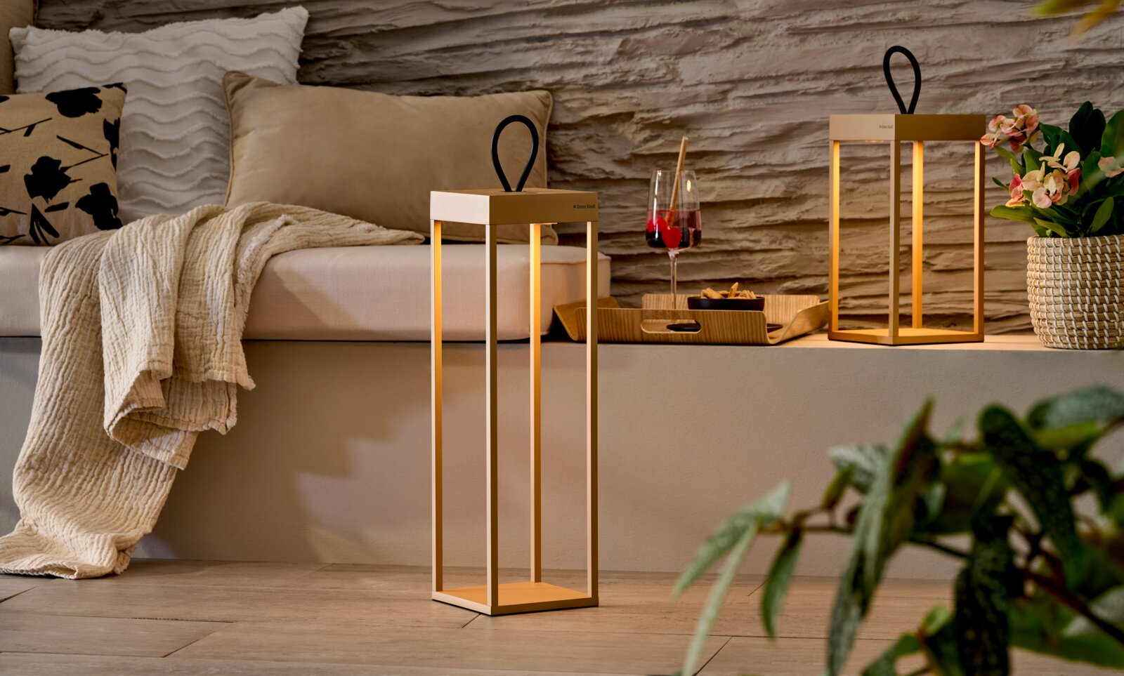 Slanke, rechthoekige led-lantaarn in goud en wit met een zwart handvat, met een warm licht, in een woonkamer op de vloer voor een bankje – perspectief: vooraanzicht op ooghoogte.