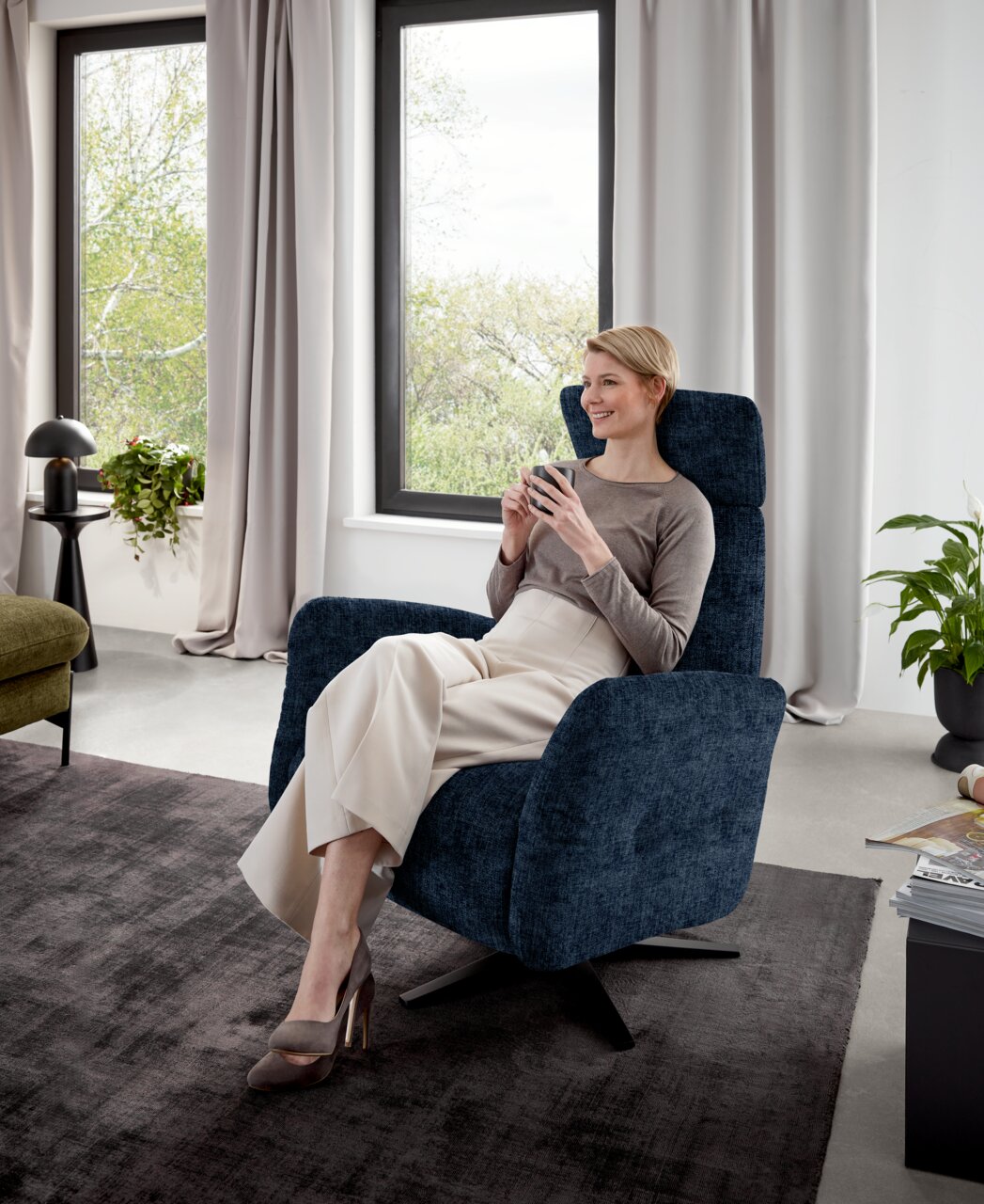 Frau sitzt entspannt in einem blauen Relaxsessel in einem modernen Wohnzimmer, seitliche Perspektive.