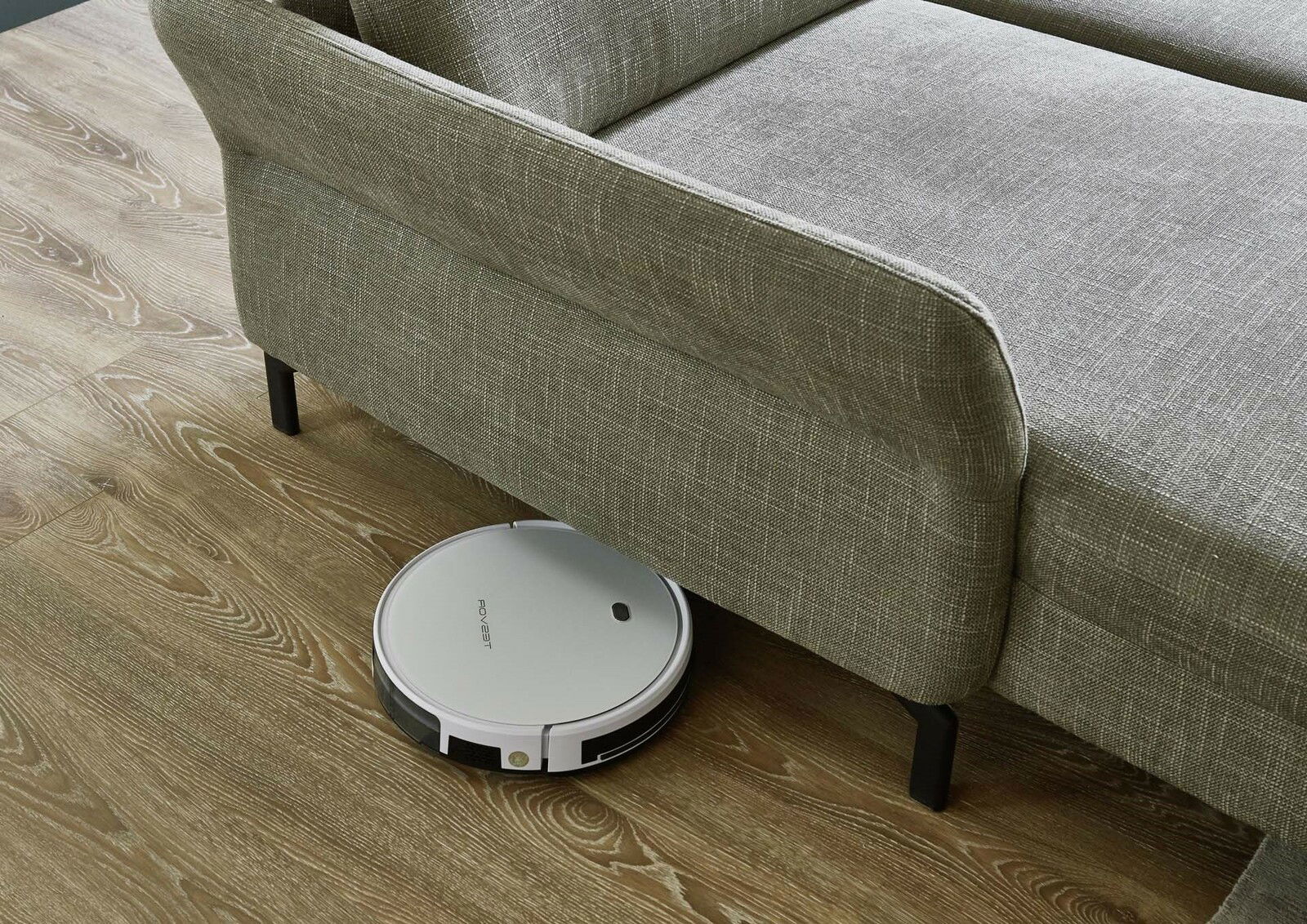 Ein graues Sofa auf einem Holzboden, darunter ein weißer Saugroboter. Perspektive von oben.