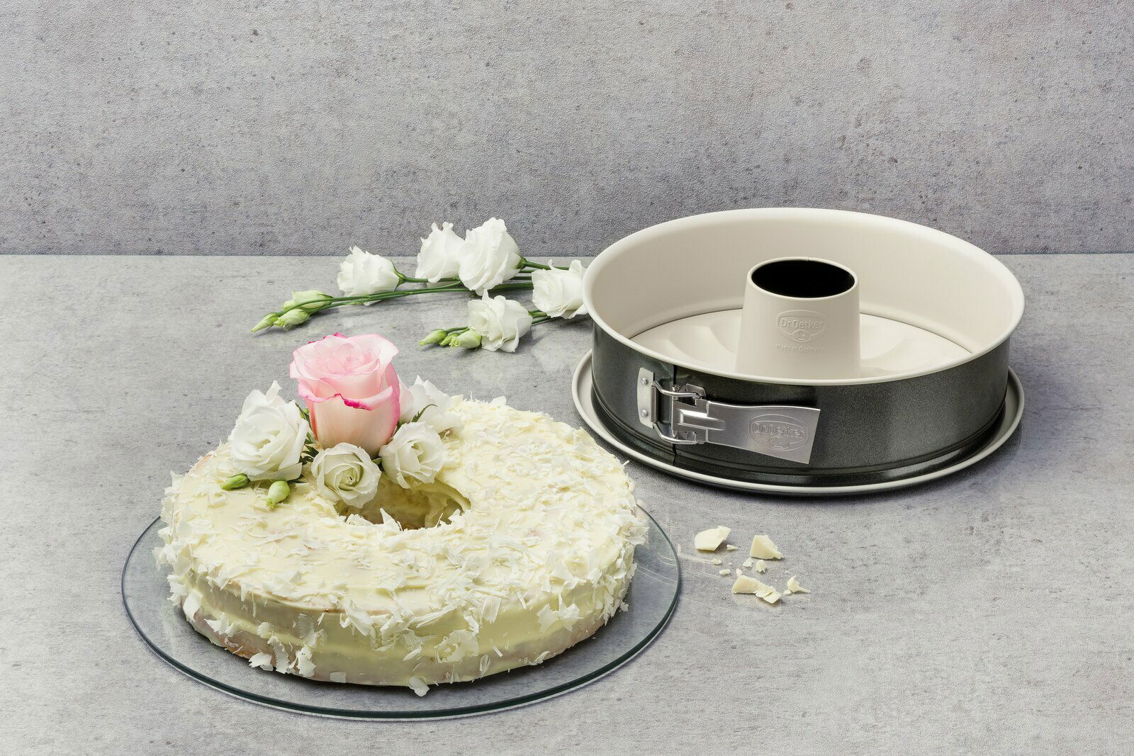 Runde Backform mit Rohrboden, 28 cm Durchmesser, neben einem dekorierten Kuchen mit weißen und rosa Blumen, aus der Vogelperspektive fotografiert.