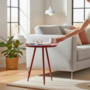 Runder Beistelltisch aus Holz in Rotbraun mit drei schlanken Beinen neben hellgrauem Sofa; darauf liegen Buch und Brille, eine Hand stellt eine Tasse ab – Perspektive: seitliche Ansicht auf Augenhöhe im Wohnzimmer.