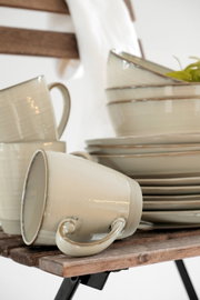 Stapel aus cremefarbenem Keramikgeschirr mit Tassen und Schalen auf Holzstuhl, eine Tasse liegt im Vordergrund seitlich; Nahaufnahme aus leicht seitlicher Perspektive